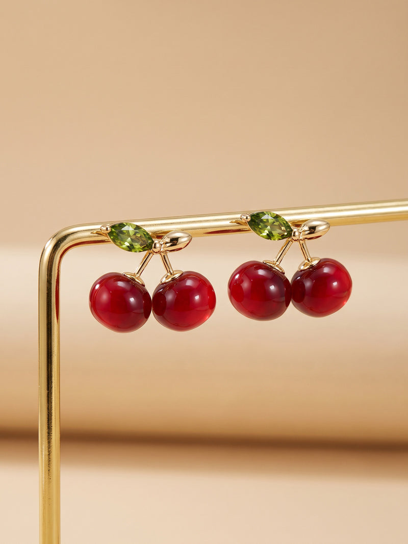 EH56: Paar roter Kirsch-Ohrstecker mit grünen Blättern an einem goldfarbenen Schmuckständer.