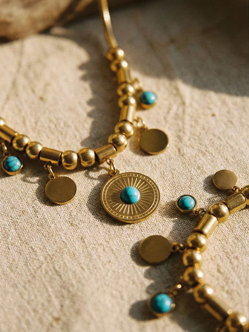 Nahaufnahme Tuerkis Sonnen Schmuck-Set auf Sand - goldener Sonnen-Anhaenger mit Tuerkis-Einlage und Muenz-Charms Detail