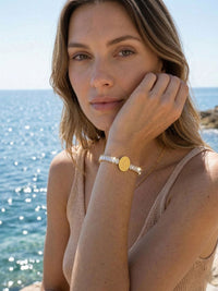 Armband Benedizione Sommer-Lifestyle – Model im Strick-Top mit Kristallarmband am Meer, Hand am Kinn, warme Sommeratmosphaere