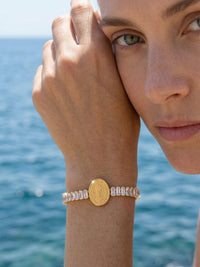 Armband Benedizione Nahaufnahme – Model haelt Hand vors Gesicht, Guadalupe-Medaillon und Baguette-Kristalle im Detail, tuerkises Meer
