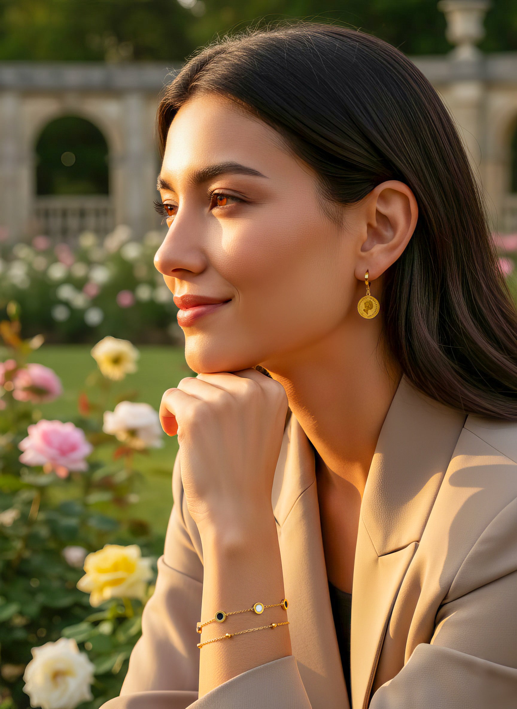 Queens Coin Ohrringe im Rosengarten – Frau mit dunklem Haar trägt vergoldete Münz-Creolen in eleganter Gartenkulisse mit warmem Sonnenlicht