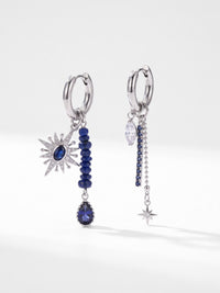 Asymmetrisches silberfarbenes Ohrringpaar mit blauen Perlen, Sonne und Stern auf hellem Untergrund mit Schatten. – EH05E