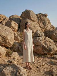 Ganzkoerper-Aufnahme Model in langem Leinenkleid mit Sommerglanz Kette vor imposanten Felsen Bohemian-Urlaubsstil