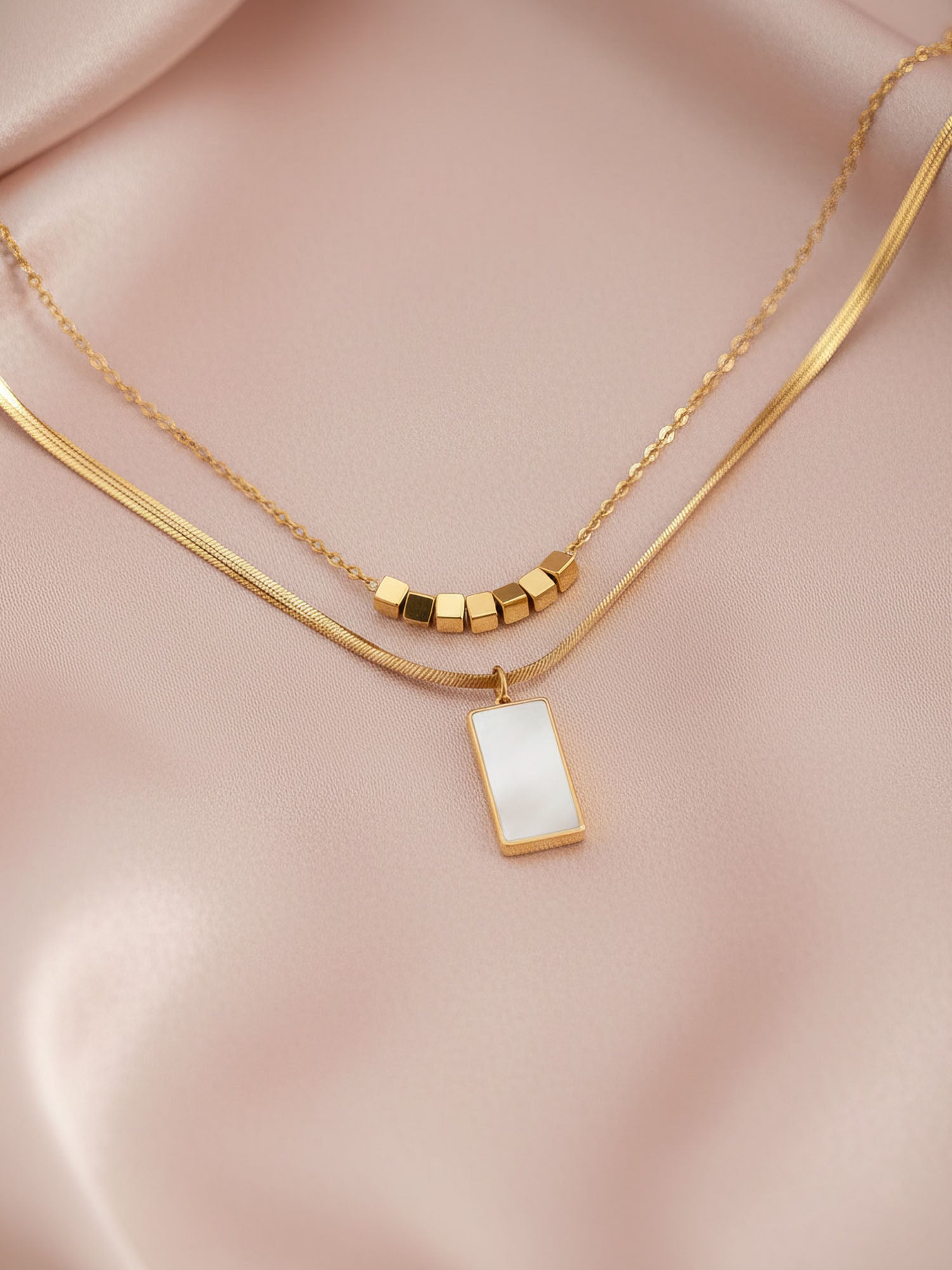 Urban Mirror Kette Flatlay auf rosa Seide – elegante Doppelkette mit Perlmutt-Anhänger und goldenen Würfelperlen