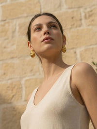 Golden Shell Ohrringe Model traegt – Frau mit erhobenem Kopf traegt vergoldete Muschel-Ohrringe, beiges V-Ausschnitt-Top, mediterrane Steinwand